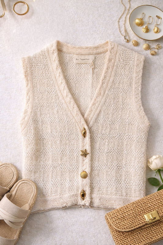 Beige knitted vest on a light surface with accessories around.  Vest has four gold seashell buttons